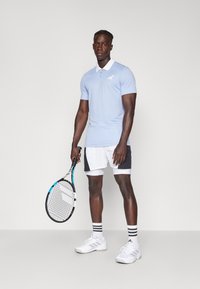 Polo de color azul claro con cuello blanco, mangas cortas y logo de Adidas. Pantalones cortos deportivos en blanco y negro, zapatillas de tenis blancas y una raqueta.