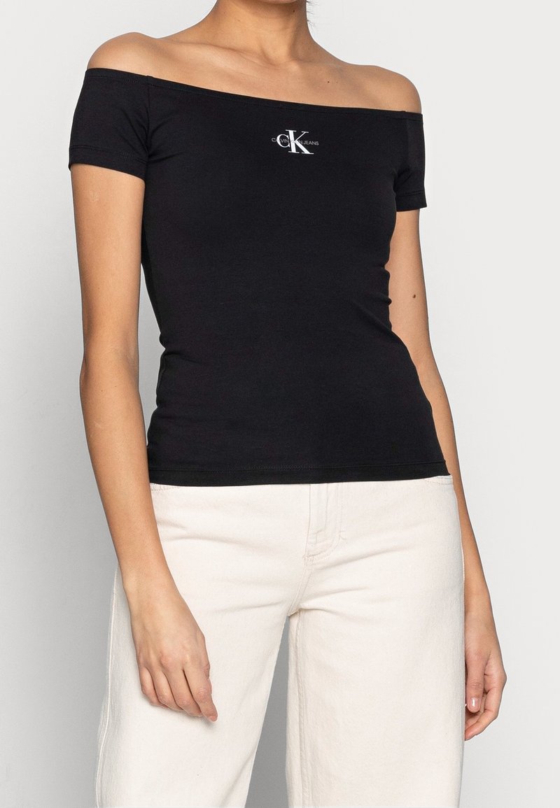 Svart off-the-shoulder åtsittande topp i bomull, med en vit logotypaccent på bröstet, tillsammans med ljusa byxor.