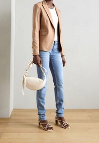 Persona vestida con una chaqueta beige, top blanco, vaqueros azul claro, sandalias de tacón beige, sosteniendo un bolso en forma de media luna color crema, de pie sobre un suelo de madera.