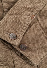 Tela de pana marrón con una superficie texturizada, que presenta dos botones de bronce grabados con "Reell". Costuras detalladas atraviesan la tela.