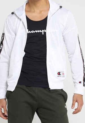Chaqueta blanca con cremallera y capucha, que presenta franjas de acento negras en las mangas y un parche de logotipo. Llevada sobre una camiseta de tirantes negra y pantalones verdes.