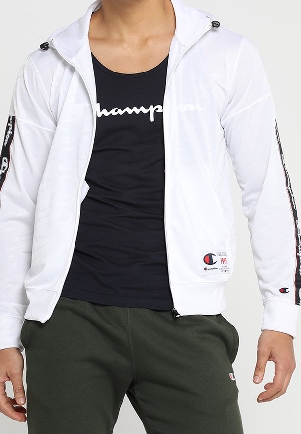 Vit zipjacka med huva, med svarta accentstripes på ärmarna och en logotypapplikation. Bärs över en svart linne och gröna byxor.