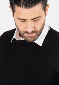 Pull en tricot noir avec un col rond, porté par-dessus une chemise blanche à col. La texture semble lisse, avec un design ajusté et sans motifs visibles.