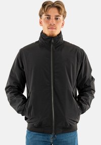 Zwarte zip-up jas met hoge kraag, elastische manchetten en zoom. Kenmerkt zich door een subtiel logo op de borst en een gladde, lichte stoftextuur.