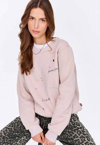 Světle růžová mikina s vyšitými frázemi v různých barvách, s dlouhými rukávy a volným střihem, doplněná leopardími kalhotami.