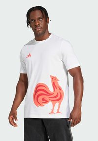 Hvid bomulds-T-shirt med et stort rødt hønegraphic på fronten, samt et lille rødt Adidas-logo nær skulderen.
