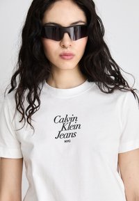 Tricou alb din bumbac cu text negru "Calvin Klein Jeans NYC". Modelul poartă ochelari de soare întunecați, cu un design unghiular și are părul ondulat.