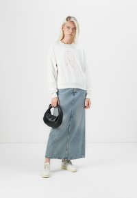 Felpa grigia chiara con grafiche a contorno rosa, abbinata a una lunga gonna di jeans blu e sneakers bianche. Tiene una piccola borsa nera.