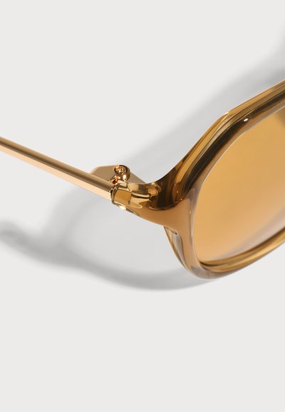 Gros plan de lunettes de soleil teintées ambre avec une monture brune translucide et des branches métalliques dorées sur fond blanc.