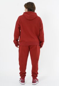Roter Hoodie und passende Jogginghosen aus weichem Stoff. Der Hoodie hat eine große, lockere Passform mit Kordelzug. Die Jogginghosen haben schmale Beine und gerippte Bündchen.