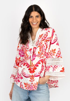 Woman smiling, wearing white blouse with red and pink floral patterns and lace trim, paired with light blue jeans, against plain background.