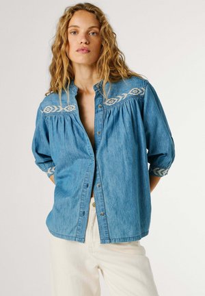 Naine laineliste blondide juustega, kandes lahtist sinist teksasärki, millel on tikitud põlled ja varrukad, koos helevärviliste pükstega, seisab valge tausta ees.