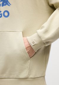 Felpa beige con tasca a marsupio, polsini a coste e logo blu. Texture liscia con dettagli di cucitura semplici.