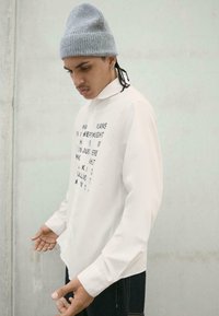 Camicia bianca a maniche lunghe con testo stampato sul davanti, abbinata a un berretto grigio in maglia. La texture appare liscia con un taglio strutturato.