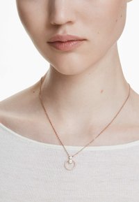 Roséguldhalsband med en cirkulär berlock, prydd med små klara stenar och en delikat kedja, bärs ovanpå en ljusfärgad topp.