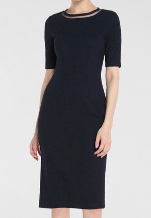Femme portant une robe noire cintrée longueur genou avec des manches courtes et une bordure noire transparente autour du décolleté.