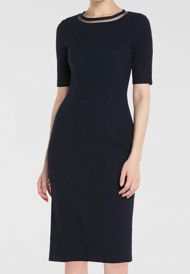 Femme portant une robe noire cintrée longueur genou avec des manches courtes et une bordure noire transparente autour du décolleté.