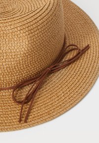 Sombrero trenzado de paja en tono marrón claro con un ala ancha. Presenta un lazo de ante marrón alrededor de la base, que ofrece un contraste de texturas y un ajuste seguro.