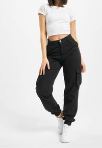 Femme portant un t-shirt blanc court, un pantalon cargo noir avec des poches, et des baskets blanches, se tenant avec une jambe légèrement fléchie.