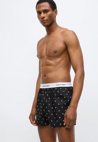 Μαύρα ανδρικά εσώρουχα τύπου μποξέρ με λευκή ελαστική ζώνη μέσης που φέρει το λογότυπο Calvin Klein και ένα επαναλαμβανόμενο διακριτικό μοτίβο από μικρά γράμματα.