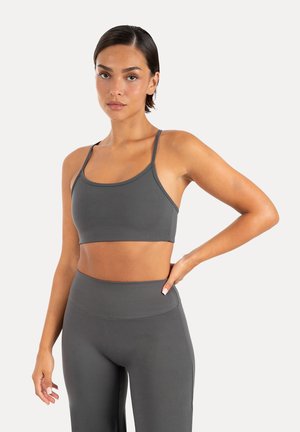 Femme aux cheveux courts et foncés portant un brassière de sport gris foncé assorti et des leggings taille haute, debout avec une main sur la hanche.