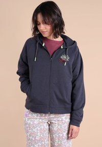 Sweat à capuche zippé gris foncé, à coupe ample, avec une capuche, des cordons de serrage verts, et une broderie florale sur la poitrine, associé à un pantalon à motifs.