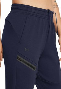 Marineblaue Jogginghosen mit einem elastischen Bund, Reißverschlusstaschen und einem kleinen schwarzen Logo an der Seite. Weicher Stoff mit glatter Textur.