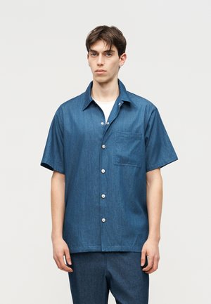 Nuori mies, joka pukeutunut siniseen, lyhythihaiseen, napilliseen paitaan, jossa on rintatasku, ja siihen sopiviin sinisiin housuihin valkoisen taustan edessä.