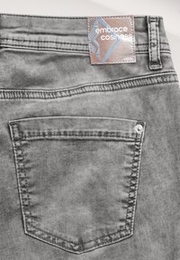Grijze denim jeans hebben een klein achterzakje, zichtbare stiksels en een label met de tekst "omarm gezelligheid" aan de tailleband bevestigd.