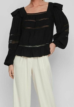 Sort bluse med lange ærmer og blondeindsatser, firkantet udskæring, flæsekant, parret med løstsiddende hvide bukser.