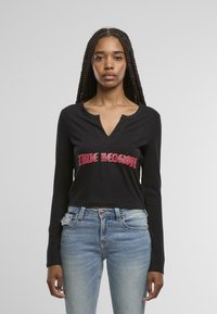 Melnas garām piedurknēm apgrieztā krekliņa ar V veida kaklu un sarkano izšūto "True Religion" logotipu priekšpusē, apvienots ar gaiši ziliem džinsiem.