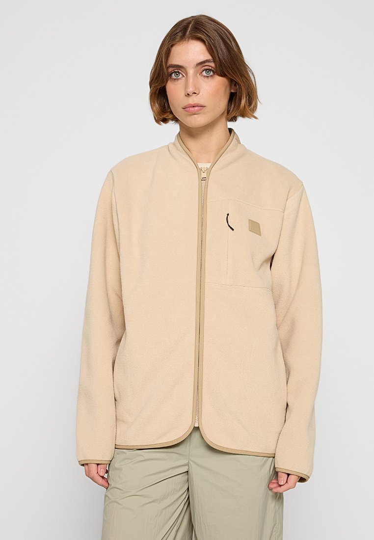 Femme aux cheveux bruns courts portant une veste en polaire beige zippée et un pantalon gris clair, se tenant devant un fond léger uni.