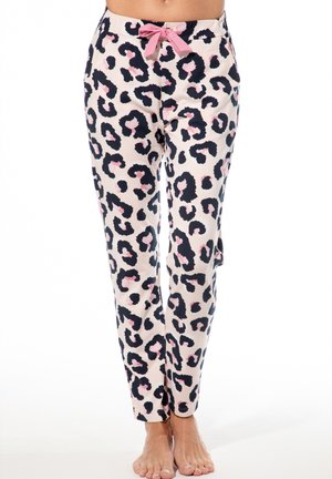 Frau, die hellfarbene Schlafanzughose mit schwarzem und pinkfarbenem Leopardenmuster und pinker Taillenbinde trägt, steht barfuß vor einem weißen Hintergrund.