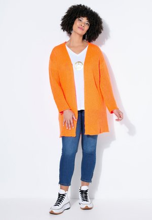 Vrouw met krullend haar, gekleed in een oranje vest, wit T-shirt met gouden vredessymbool, blauwe spijkerbroek en witte hoge sneakers, staat ontspannen.