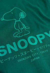 T-shirt en coton vert avec un graphisme de Snoopy et du texte en plusieurs langues, dont « SNOOPY » et « SHINJUKU CITY, TOKYO, JAPAN ».