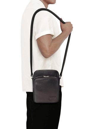 Personne portant une chemise blanche à manches courtes porte un petit sac bandoulière en cuir noir avec une poche avant zippée.