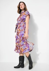Lila gemustertes Kleid mit kurzen Ärmeln, tailliertem Schnitt und ausgestelltem Rock. Kombiniert mit schwarzen Leder-Cowboystiefeln.