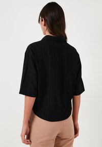 Black, ribbed short-sleeve blouse with a collar and a rounded hem. Worn over beige pants. Smooth texture, casual fit, and subtle design.