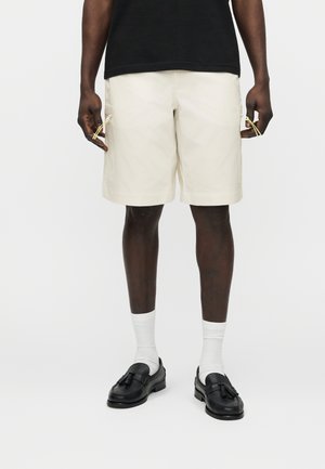 Άνδρας με κρεμ cargo σορτς με πλαϊνές τσέπες, λευκές κάλτσες μέχρι τον άστραγα, μαύρα loafers με φούντες και μαύρο t-shirt σε ένα απλό λευκό φόντο.