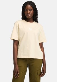 Béžové tričko s krátkým rukávem vyrobené z bavlny s čtvercovým logem na hrudi. Tričko má uvolněný střih a hladkou texturu.