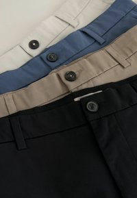 Quatre paires de pantalons pliés superposées, montrant des ceintures avec boutons, passants de ceinture et couleurs : noir, beige, bleu et écru.