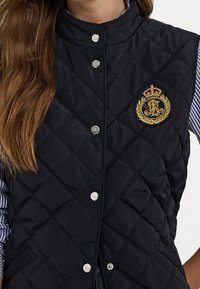 Vadderad svart väst med silvriga tryckknappar. Har en broderad gyllene krona med lagerkransdetaljer på bröstet.