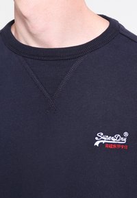 Primo piano di una persona che indossa un felpa SuperDry blu navy con colletto a coste e logo ricamato sul petto.