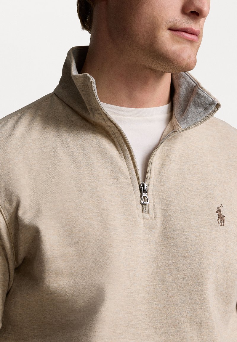 Pullover beige a zip con mezze maniche realizzato in materiale morbido, con colletto interno grigio a contrasto e piccolo logo ricamato sul petto.