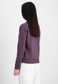 Lilla sweatshirt med ribbede manchetter og kant, lavet af blødt stof. Modellen har hvide brede bukser på og står med ryggen til kameraet.