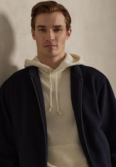 Tumma laivastonsininen takki kerrospukeutumisessa käytetyn kermanvärisen, teksturoidun neulottu hupullisen paidan päällä, jossa on kiristysnauha. Pehmeät materiaalit ja rento, kerrostettu muotoilu.