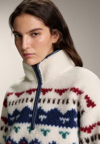 Fleece-tröja med hög krage, med en vit bas och färgglada geometriska mönster i rött, blått och grönt. Blixtlås och texturerat tyg.