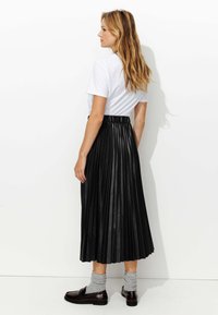 Jupe midi plissée noire en matériau brillant, portée avec un t-shirt blanc, des chaussettes grises et des chaussures sombres. La taille est élastique.