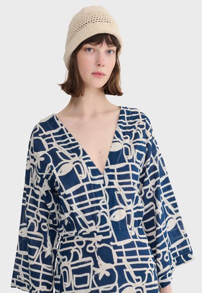 Młoda kobieta z krótkimi brązowymi włosami, ubrana w beżową dzianinową czapkę i niebieską sukienkę z białym abstrakcyjnym geometrycznym wzorem.