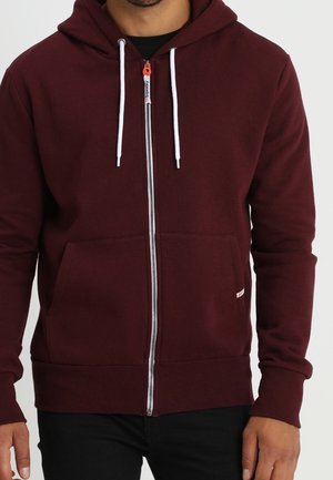 Mann trägt ein burgunderrotes Kapuzen-Sweatshirt mit Reißverschluss, weißen Kordeln und Fronttaschen, kombiniert mit schwarzen Hosen.